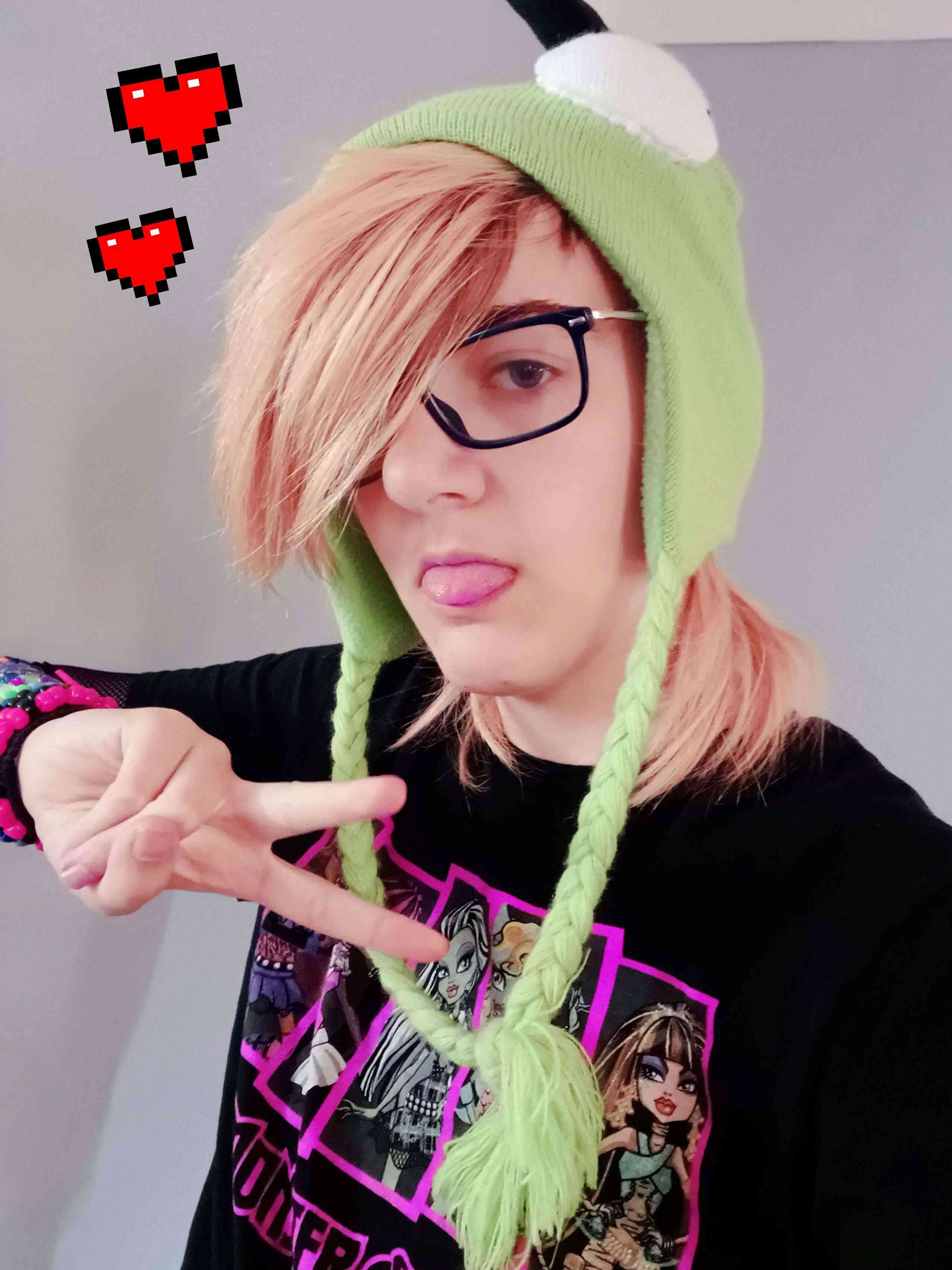 a boy wearing a green hat and a t-shirt with monster high characters. he has bleached hair, glasses, and is holding up a peace sign while sticking out his tongue.